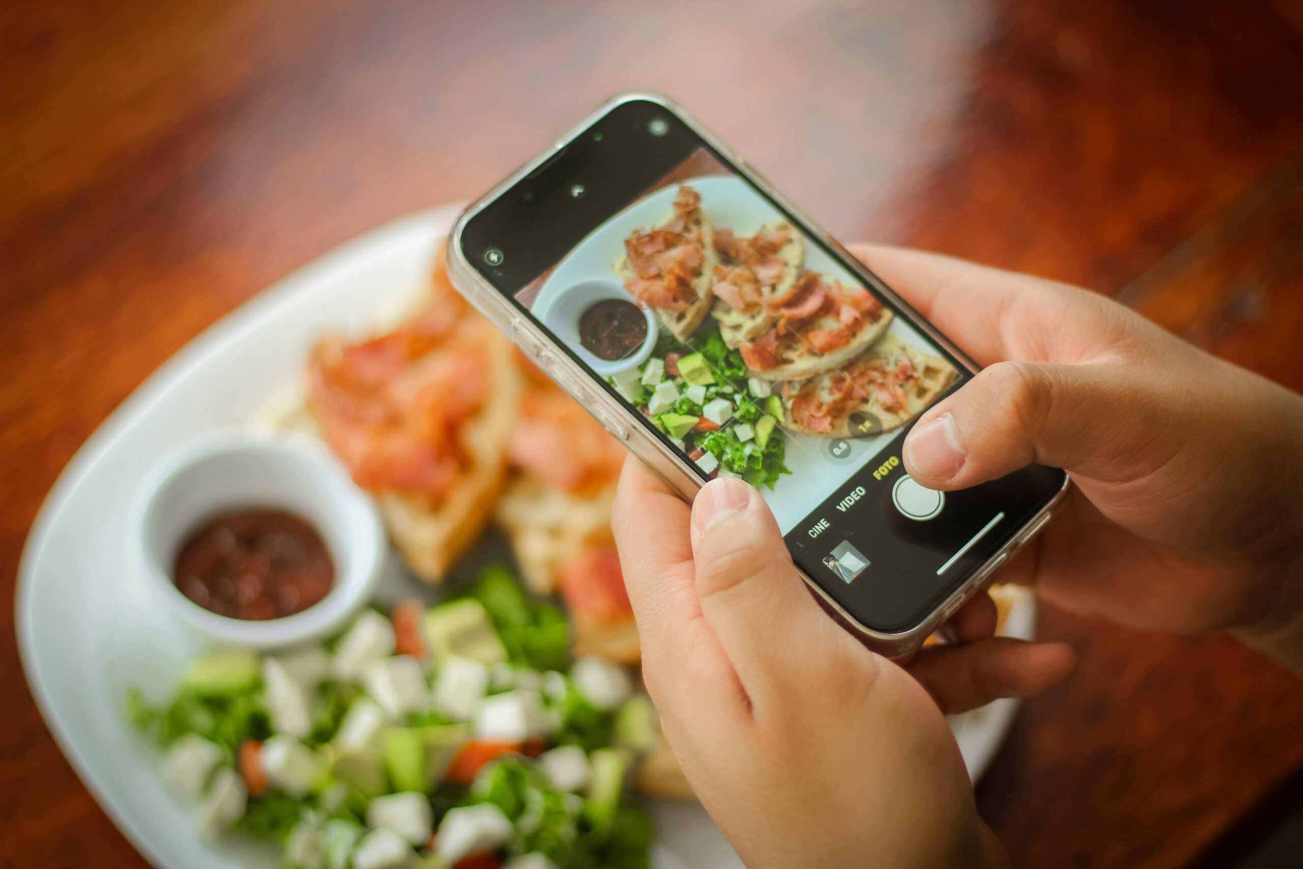 Nahaufnahme von HÃ¤nden, die mit einem Smartphone einen Brunchteller mit Speck, Salat und SoÃŸe fotografieren.